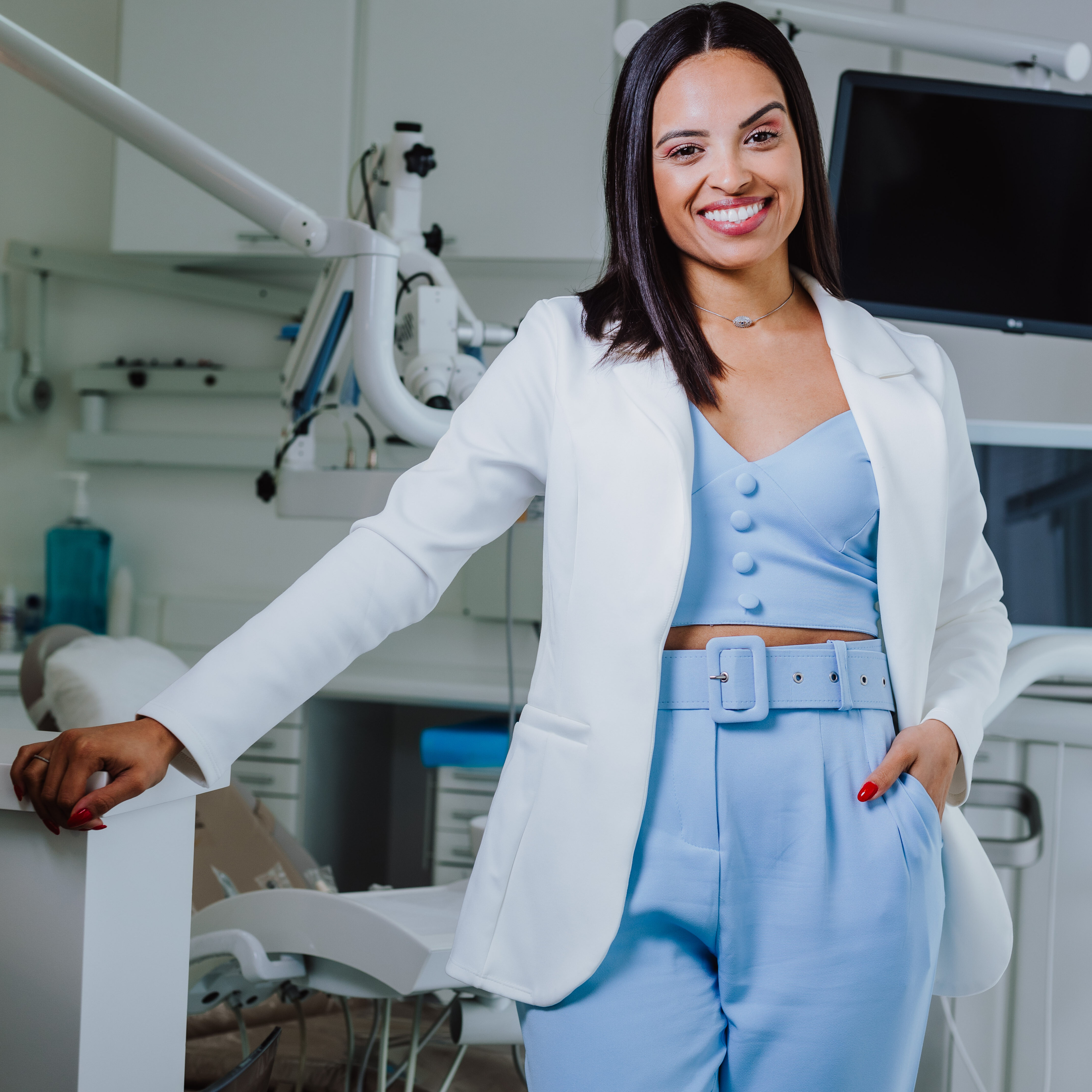 Dentista sorridente em consultório odontológico moderno. Uma dentista jovem e profissional, vestindo um conjunto elegante azul claro com blazer branco, está em pé ao lado de uma cadeira odontológica em um consultório moderno e bem equipado. Ela transmite confiança e simpatia, pronta para oferecer atendimento de qualidade aos pacientes. Ambiente claro e acolhedor, ideal para tratamentos dentários como limpeza, clareamento, implantes dentários e outros procedimentos de odontologia estética.
